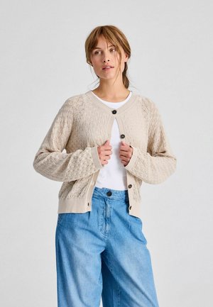 Cardigan en maille beige avec des motifs en losanges, col rond et quatre boutons noirs. Associé à une chemise blanche et un jean bleu clair.