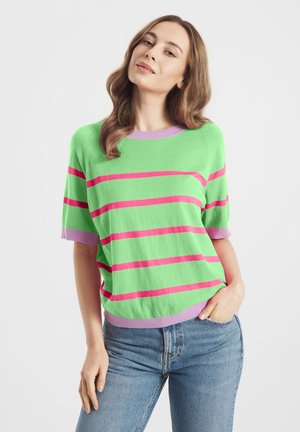 Vrouw met golvend bruin haar, gekleed in een groen kortemouwentrui met roze strepen en lila rand, en een blauwe spijkerbroek, staand met één hand in haar zak.