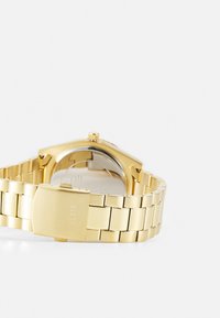 Montre en métal doré avec un bracelet à maillons, dotée d'un boîtier lisse, d'une couronne et d'un fermoir avec le logo "GUESS" gravé.
