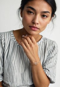 Silverarmband med ett cirkulärt träddesign och små pärlor, bärs på en modells handled. Modellen har lätt, randig tyg med rund ringning.