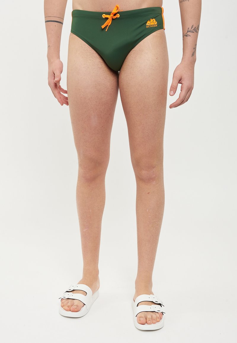 Un slip de bain vert foncé avec un cordon de serrage orange, arborant un logo sur le côté. Porté avec des sandales glissantes blanches. Matériau lisse et design ajusté.