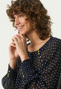 Blouse bleu marine à pois, en tissu transparent, avec des poignets smockés et des boutons dorés le long du col, créant un look texturé et superposé.