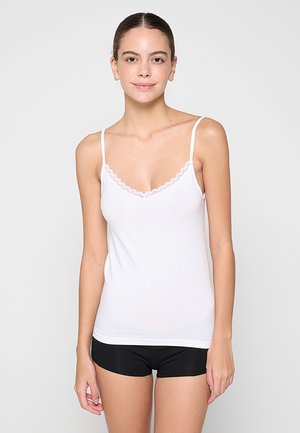 Junge Frau, die ein weißes Camisole-Top mit Spitzenbesatz und schwarze Shorts trägt, steht vor einem schlichten hellgrauen Hintergrund.