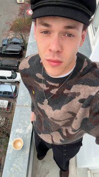 Pletený svetr v hnědém, černém a šedém maskáčovém vzoru, nošený s tmavými kalhotami. Viditelné jsou stříbrné náhrdelníky a světle hnědý hrnek na kávu.