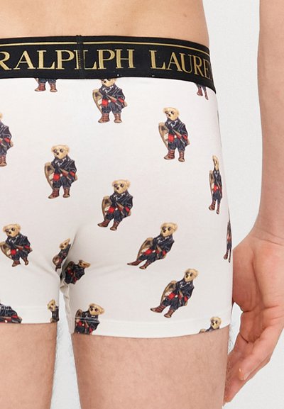 Biele boxerky s opakujúcim sa vzorom oblečeného medveďa. Čierny pás so zlatým nápisom "RALPH LAUREN".