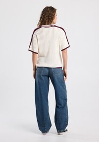 Knit oversized sweater in cream with maroon accents, featuring a ribbed collar and short sleeves, paired with loose blue jeans.