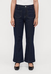 Kvinna i mörkblå utsvängda jeans med framfickor och bruna Mary Jane-skor, stående mot en enkel vit bakgrund.