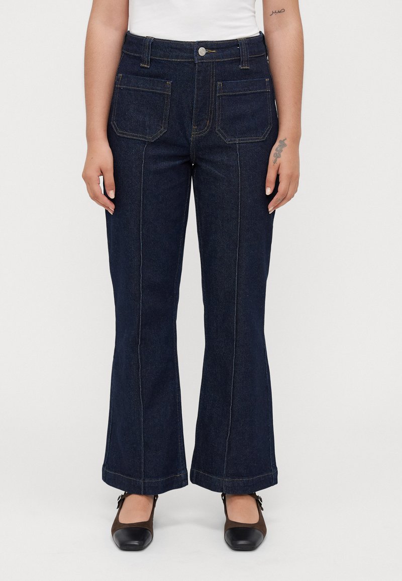 Kvinna i mörkblå utsvängda jeans med framfickor och bruna Mary Jane-skor, stående mot en enkel vit bakgrund.