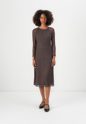 Robe marron à manches longues, transparente, avec un motif à pois et un ourlet festonné, fabriquée en tissu léger. Associée à des chaussures noires à talons plats.