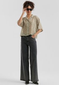 Camicia trasparente di un leggero verde oliva con pois bianchi, maniche corte e colletto, abbinata a jeans a vita alta grigio scuro con gamba larga. Occhiali da sole neri.