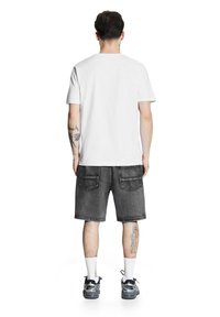 Biała koszulka z krótkim rękawem w połączeniu z czarnymi szortami dżinsowymi. Buty są szare z czarnymi akcentami. Widoczne tatuaże na lewej ręce.