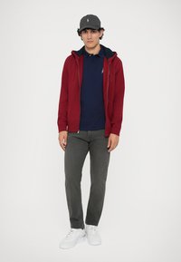 Sweatshirt vermelha com fecho e bolsos, camisa polo azul-marinho por baixo, calças cinzentas e ténis brancos. Boné cinzento com logótipo completa o conjunto.