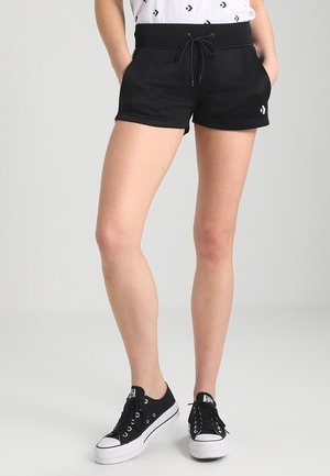 Femme portant un short noir à cordon avec des poches latérales et des baskets noires basses, debout les mains dans les poches sur un fond gris.