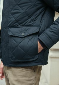 Marineblau gesteppte Jacke mit einer Klappen-Tasche und Druckknopf, strukturiertem Stoff und glatter Oberfläche, kombiniert mit hellbeigen Hosen.