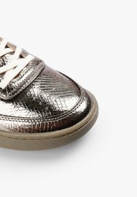 Zapatilla plateada metálica con patrón en relieve, cordones blancos y suela de goma texturizada. Presenta perforaciones en la puntera.