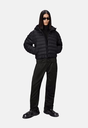 Persona che indossa una giacca imbottita nera, pantaloni neri, scarpe nere e occhiali da sole rettangolari neri, in piedi con le mani nelle tasche della giacca.