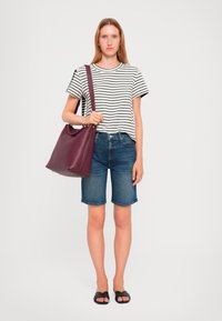 T-shirt a righe a maniche corte, pantaloni shorts in denim blu, sandali neri, tiene in mano una borsa in pelle bordeaux dal design morbido con dettagli in metallo dorato.