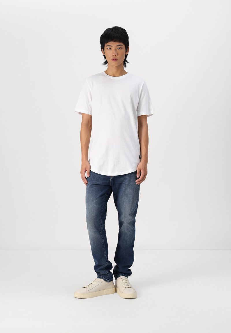 T-shirt blanc à col rond, jean en denim bleu et baskets beiges. Le t-shirt a une coupe décontractée ; les jeans présentent une légère décoloration et les baskets ont une semelle épaisse.
