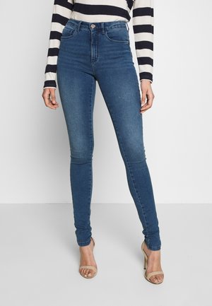 Femme portant un jean skinny bleu taille haute, un haut à manches longues rayé et des sandales beiges à talons ouverts, debout sur un fond uni.