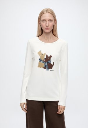 Femme blonde portant une chemise blanche à manches longues avec des chiens dessinés portant des écharpes, associée à un pantalon marron foncé sur un fond uni.