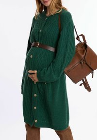 Groene gebreide cardigan met kabelpatroon, lange mouwen, sluitingen met knoppen en een bruine riem. Vergezeld van een bruine lederen rugzak.