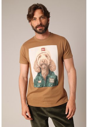 Man with beard wearing brown t-shirt featuring a dog in a beanie and jacket with "Born Chill" patch.