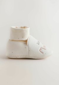 Soft cream-colored baby bootie with folded cuff and subtle dotted pattern, lying on a plain light background.