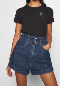 Mujer vestida con una camiseta negra con el texto verde "FLWR PWRS" y pantalones cortos de mezclilla azul oscuro de talle alto con los puños doblados, con una mano en el bolsillo.