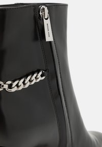 Bottine en cuir noir avec une fermeture éclair sur le côté, dotée d'une tirette métallique et d'un accent de chaîne argentée sur le côté. Texture lisse, design épuré.