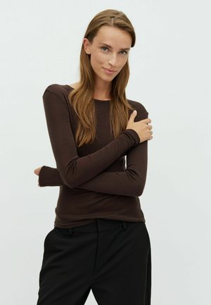 Donna con capelli castani lunghi che indossa una maglia aderente marrone scuro a maniche lunghe e pantaloni neri, in piedi con le braccia incrociate e un leggero sorriso.