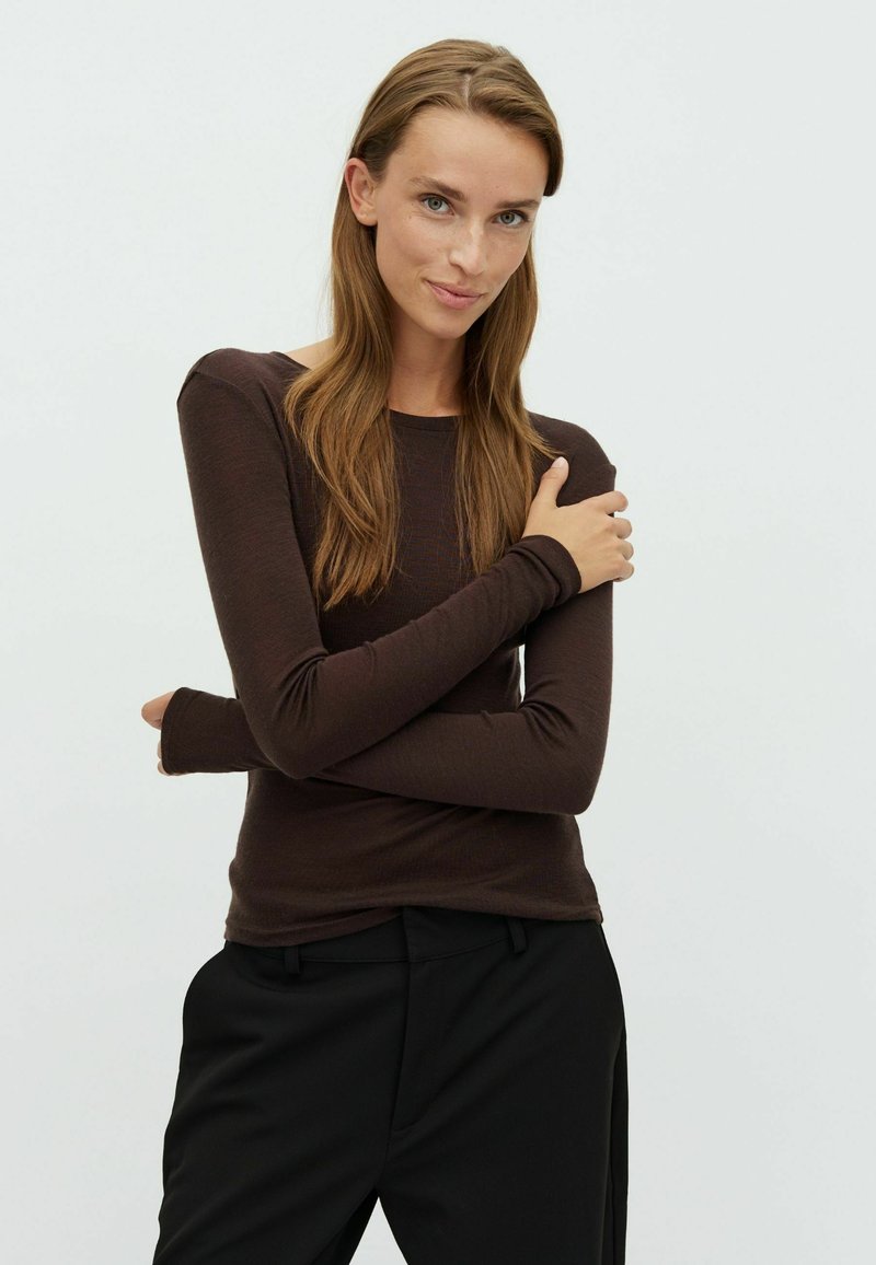 Femme aux longs cheveux bruns portant un haut à manches longues marron foncé ajusté et un pantalon noir, debout les bras croisés avec un léger sourire.