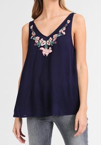 A model wears a navy sleeveless top featuring floral embroidery around the neckline, paired with light jeans.