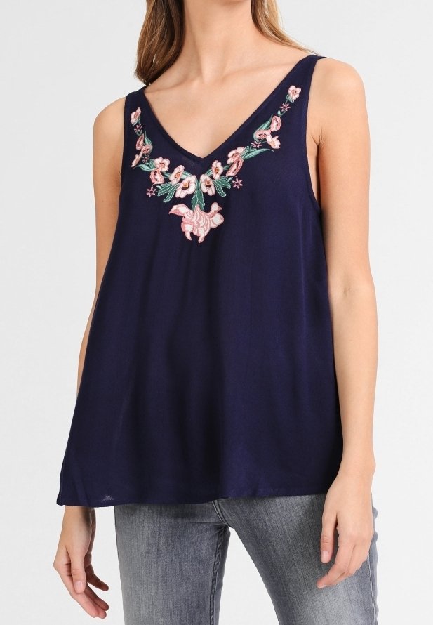 A model wears a navy sleeveless top featuring floral embroidery around the neckline, paired with light jeans.