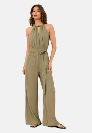 Kobieta ubrana w oliwkowo-zielony bezrękawnikowy kombinezon z szerokimi nogawkami i przewiązanym paskiem w talii, pozująca z jedną ręką w kieszeni i sandałach.