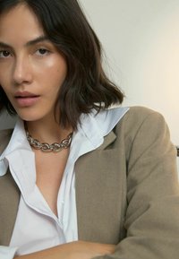 Silberne Kettenhalskette an einer Person, die einen beigen Blazer und ein weißes, kragenloses Hemd trägt. Das Oberteil ist geschichtet, mit sichtbarer Gewebestruktur.