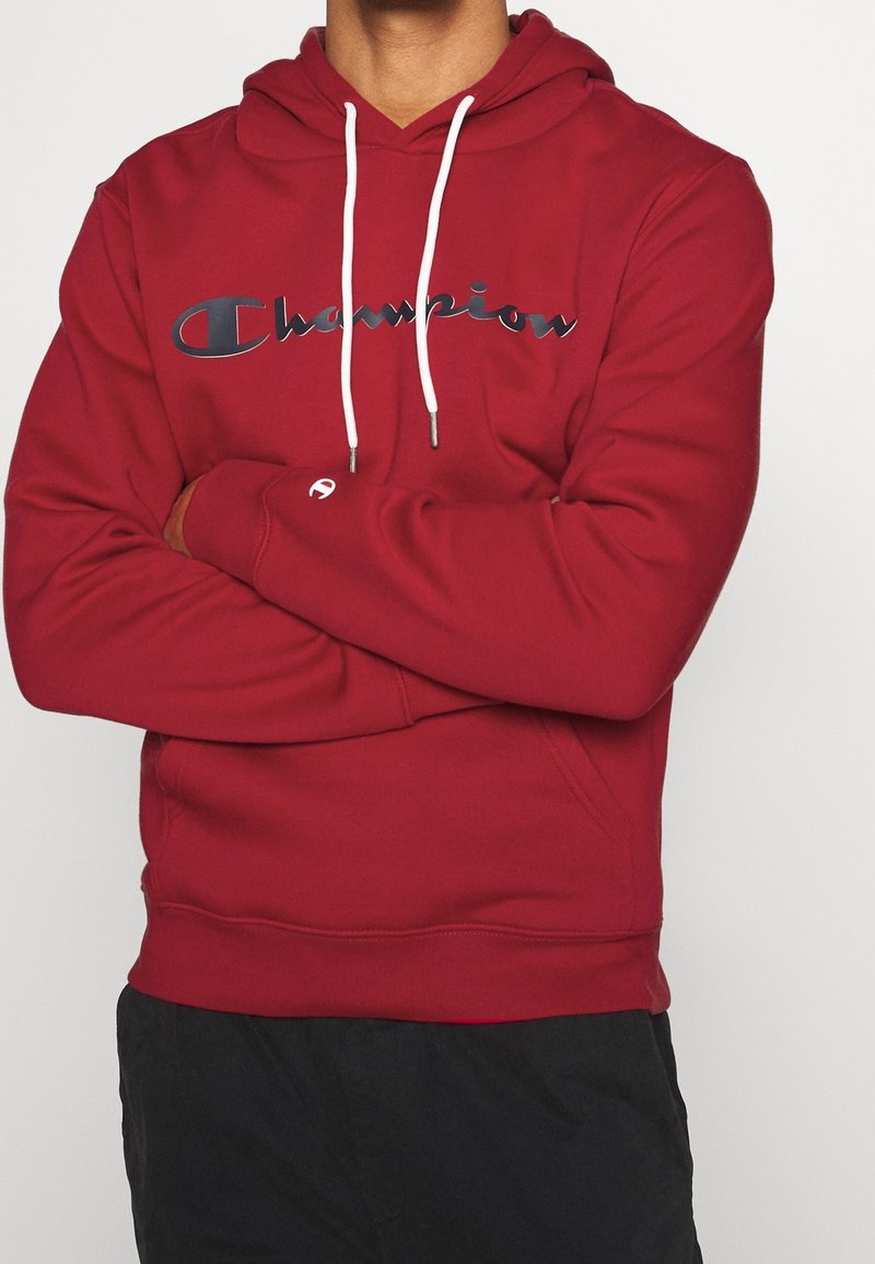 Sweat à capuche rouge en tissu mélangé coton, présentant un logo "Champion" bien visible en bleu foncé, des cordons blancs et des poignets côtelés.