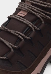 Κοντινή λήψη σκούρου καφέ αθλητικού παπουτσιού με κορδόνια σε γκρι ζιγκ-ζαγκ μοτίβο και επενδεδυμένη γλώσσα με ροζ υφασμάτινη θηλιά.