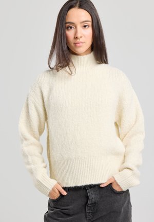 Maglione a collo alto crema con texture a coste, vestibilità rilassata e maniche lunghe. Abbinato a pantaloni alti grigi scuri.