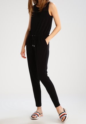 Zwarte mouwloze jumpsuit met een elastische tailleband en verstelbare trekkoord. Gecombineerd met witte slippers met zwarte, rode en blauwe strepen.