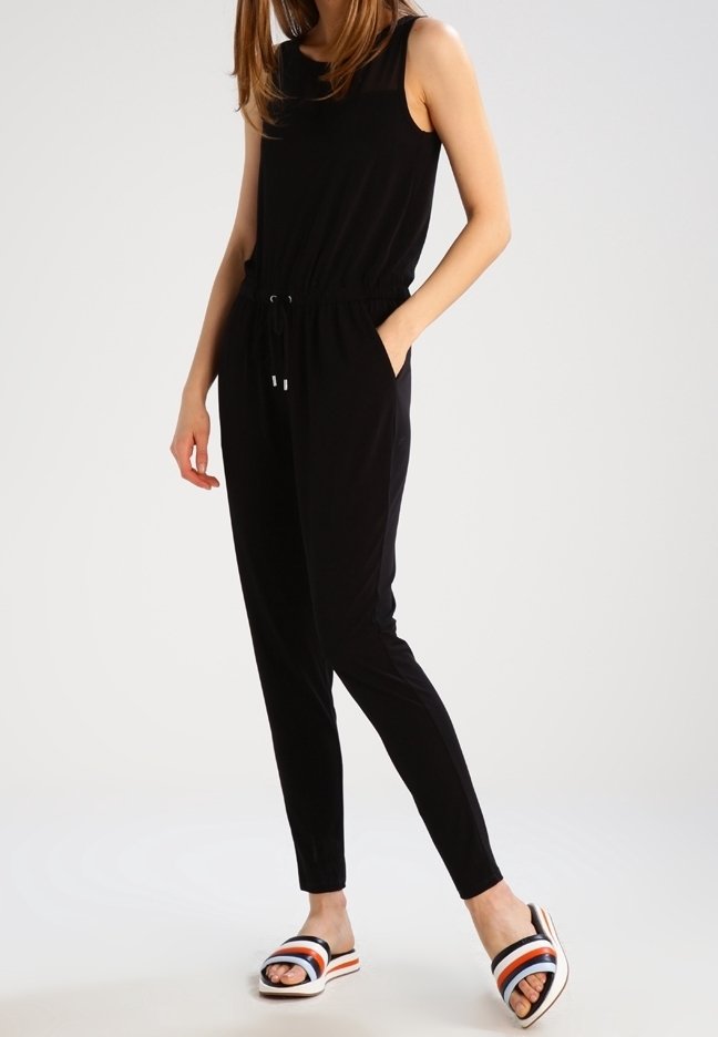 Combinaison noire sans manches avec une taille élastique et un cordon ajustable. Associée à des sandales blanches avec des rayures noires, rouges et bleues.