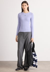 Hellblau-langes Oberteil mit Rippenstruktur, kombiniert mit grauen, gestreiften weiten Hosen. Schwarze flache Schuhe und eine schwarze Tragetasche.