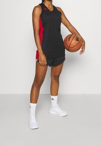 Uniforme de baloncesto negro y rojo con una camiseta sin mangas, combinado con pantalones cortos negros, calcetines blancos y zapatillas blancas, sosteniendo un balón de baloncesto.