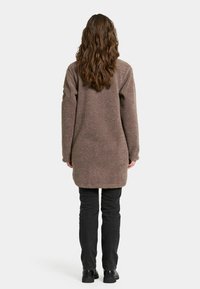 Lang brun fleece frakke med en tekstureret overflade, lige snit, sænkede skuldre og sidelommer, parret med sorte tætsiddende bukser og støvler.