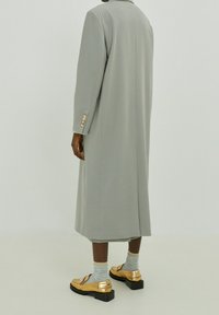 Long manteau gris à la coupe droite, texture lisse et boutons dorés en accent sur les manches. Associé à des chaussures dorées brillantes et des chaussettes rayées.