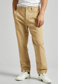Chinos de algodón beige con un corte recto, que cuentan con dos bolsillos laterales y un cierre frontal con botones. Combinados con zapatillas blancas.