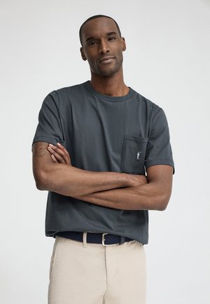 Mężczyzna z założonymi ramionami, ubrany w ciemnoszarą koszulkę z kieszeniami i beżowe spodnie na jednolitym jasnym tle.