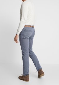 Pánské modré vzorované kalhoty se slim fit střihem; mají hnědý pásek, postranní kapsy a jsou doplněny hnědými semišovými botami.