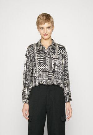 Camisa de patrón en blanco y negro con mangas largas, cuello abotonado y diseños geométricos y florales intrincados. Combinada con pantalones negros.