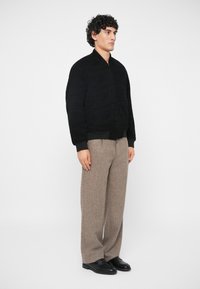 Άνδρας με σγουρά μαλλιά που φοράει ένα μαύρο μπόμπερ τζάκετ, μπεζ παντελόνια με φαρδιές γάμπες και μαύρα παπούτσια, στέκεται μπροστά από ένα απλό λευκό φόντο.