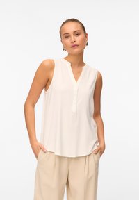Blusa senza maniche di colore crema con colletto a banda, chiusura frontale a bottoni e tessuto leggero e fluido. Abbinata a pantaloni beige larghi.